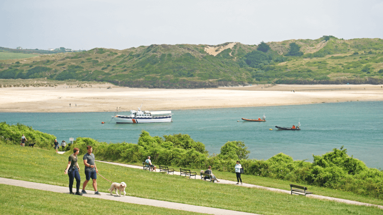 People wander on accessible paved paths along the coastline on the Camel Trail, with the estuary in the background.