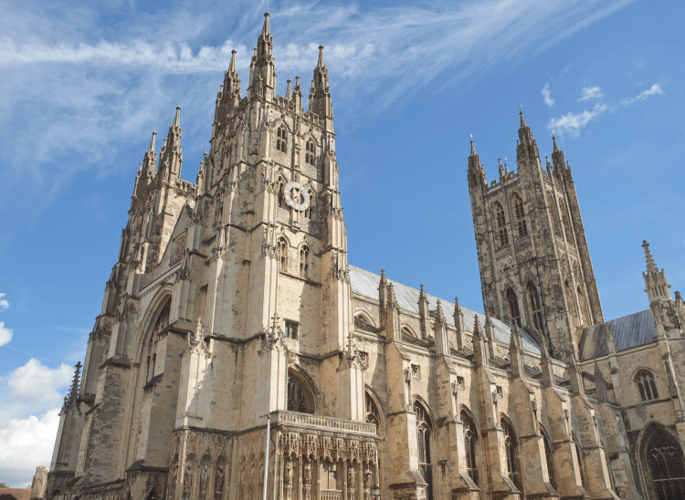 An upward view of Canterbury Cathedral on the Old Way pilgrimage route. It's an impressive cathedral with three square towers, a fine leaded roof, supporting buttresses and a host of ornate windows and carved details, all in pale stone.