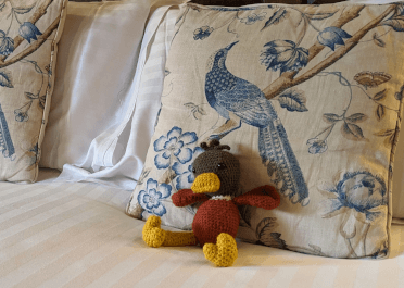 Contours Holidays' crocheted duck mascot sits on the bed, relaxing against decorative cushions embroidered with peacocks.