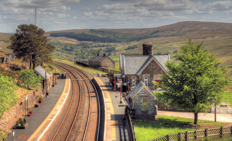 A high view of Dent Station, a square stone building with gabled roof that sits alongside a curving railway track. Green fields lie on all sides.