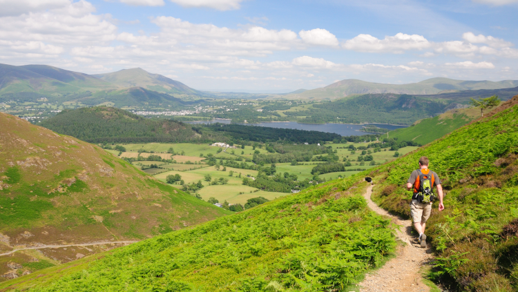 A walker in summer kit descends a footpath towards a lake and distant hills.