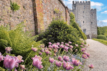 A red stone wall lined with tulips leads the eye through a manicured garden toward the square-walled pele tower of Sizergh Castle.