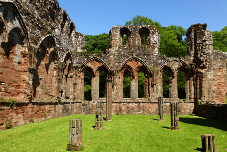 A view from within the ruin of Furness Abbey, a roofless red-stone building with ornate arched windows and several old stone pillars that have since crumbled through time.