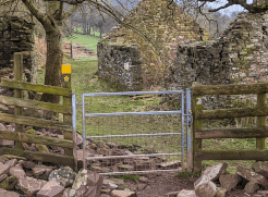 A galvanised pedestrian gate, marked with a right-of-way fingerpost, brings the path between two ruined stone farm buildings and toward the fields beyond.