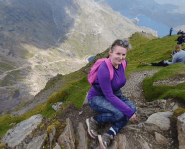 Gosia sits near the cliff edge towards the top of Mount Snowdon. The ground drops away steeply behind her to rocky mountain slopes and a lake below.