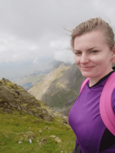 Gosia poses for a selfie at the summit of Snowdon, with windswept views of the mountain slope behind her.