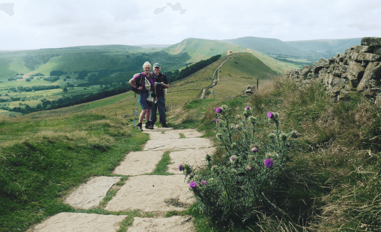 A couple stand on the Great Ridge Walk in hiking gear, midway through the Mam Tor Circular Walk. The path is set in broad flagstones in the foreground, and in the background, the ridge of the hill marches ahead toward Mam Tor with steeply sided valleys dropping away on either side.