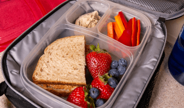 An insulated lunchbox sits open to display its contents: hummus, dipping vegetables, a sandwich, strawberries and blueberries. This type of lunchbox is great for keeping food cool on the trail.