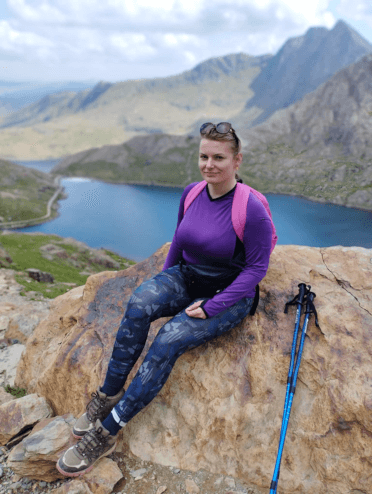 In walking gear with walking poles, Gosia leans against a large rock to rest on her descent down Snowdon's Miners' Track. Behind her, we see other rocky pecks and a bright blue lake.