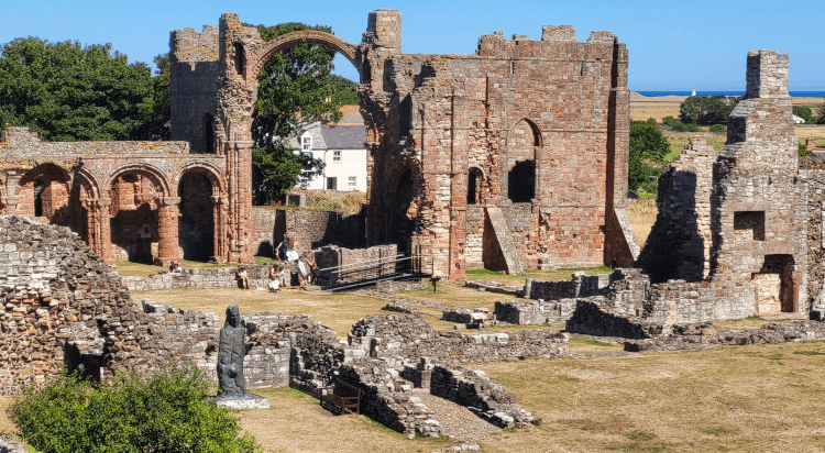 An elevated view through into the ruin of Lindisfarne Priory on St Cuthberts Way, one of our most popular pilgrim routes in the UK. None of the walls are complete, but several ornate arches remain, including an impressive two-storey arch. A statue of St Cuthbert himself stands just outside the walls.