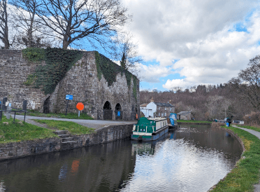 A photo of the modern-day restored remains of the Llangattock Lime Kilns, a large stone building with inset arches that stands slightly set back from the canal, with a boat moored nearby.