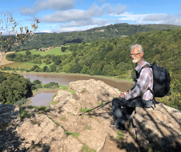 A walker dressed in loose, protective clothing for summer sits on a rock overlooking a curving stretch of river running through the valley below.