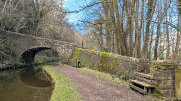 The Usk Valley Walk follows the towpath of the Monmouthshire and Brecon Canal, which here is a secluded, well-surfaced pathway that follows the curve of the canal beneath a low packhorse-style bridge flanked by woodland.