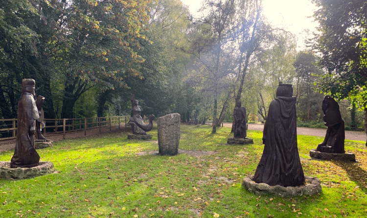 A series of statues surround a standing stone in the middle of a field, with autumn sunlight streaming down over the scene.