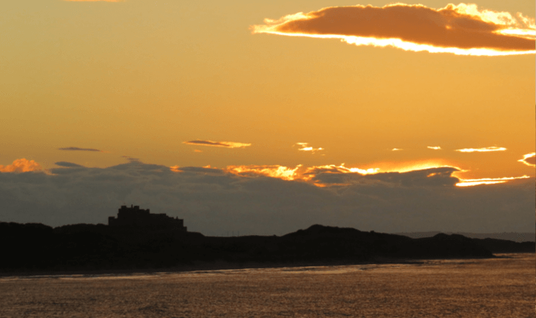 A yellow autumn sunset behind the black silhouette of a seaside castle on the Northumberland Coast Path
