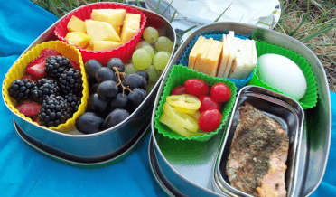 Food to be carried on a walking trail, held securely in two metal lunchboxes and divided up with silicon dishes. The left lunchbox is full of grapes, berries and cheese, while the right lunchbox contains cheese, an egg, salmon, plum tomatoes and peppers.