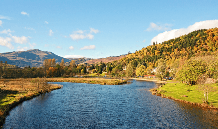 A broad stretch of river on the Rob Roy Way, with trees on either side and the bulk of a Scottish mountain in the distance.