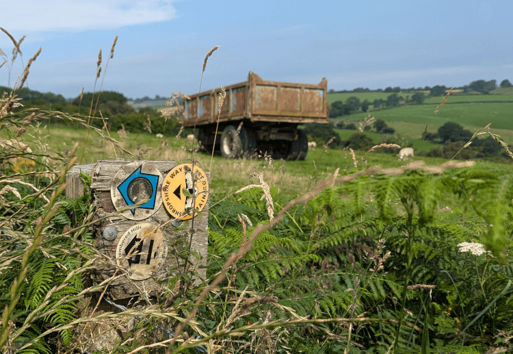 A Shropshire Way waymark stands from the hedge at a field boundary, with a large farming trailer parked in the field behind.