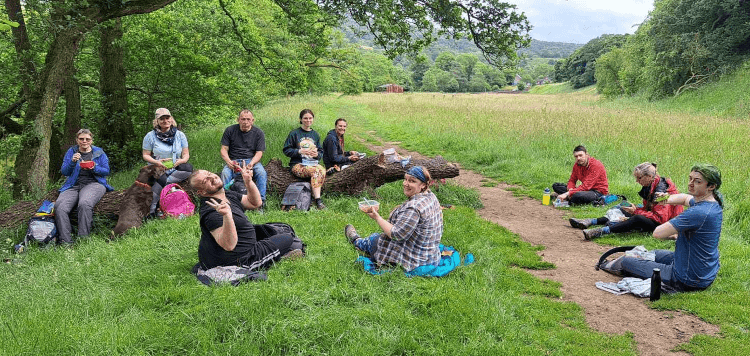 A group of walkers relax under a tree as they eat packed lunch midway through a summer walk.