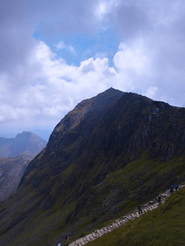 A view from far up Mount Snowdon towards the rocky crag that forms the summit. It is a huge, dark, rocky protrusion with a bright blue sky behind.