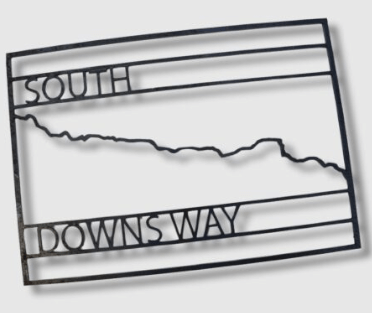 A metal trail sign depicting the South Downs Way. The name of the trail is captured in block caps, with the twisting line of the trail captured in the middle.