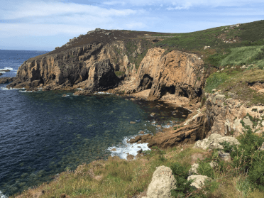 The South West Coast Path clambers along the clifftops above this rocky coastal cove, with the sea below, as bright as the blue summer sky overhead.