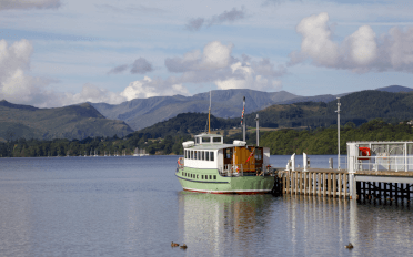 On a clear spring day, a green passenger craft is moored at a wooden pier on Ullswater, with the mountains of the Lake District in the distance.