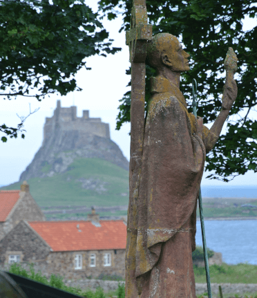 A statue of St Cuthbert stands side-on in the foreground. Past him, through overhanging leaves and over a red-roofed house, Holy Island and Lindisfarne Castle stand in the sea. This stunning view can be found on the Northern Saints Trails, the Forth to Farne Way, and St Cuthbert's Way.