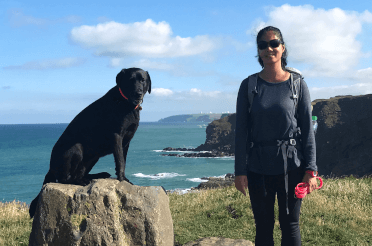 A walker poses on a coastal path with the sea beside her and her dog sitting on a large rock beside her. She wears large-lensed sunglasses to protect her eyes.