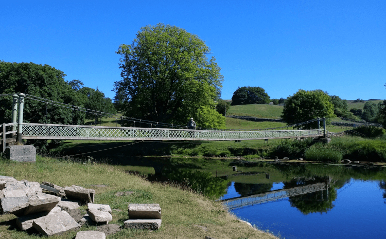 A narrow pedestrian suspension bridge spans the River Wharfe on the Dales Way. The bridge is remarkably long, with the blue waters of the river below and a blue sky above, with green hills and plenty of trees all around.