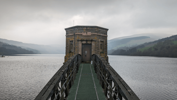 A view along the metal walkway to the Talybont Reservoir Valve Tower, a hexagonal stone tower rising from the water.