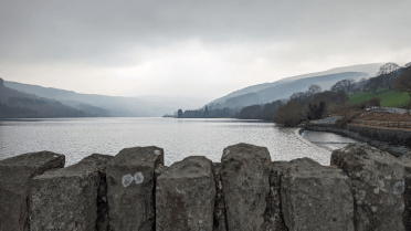 A view over the topping stones of a stone wall over the Talybont Reservoir, an expanse of still water surrounded by steep hills.