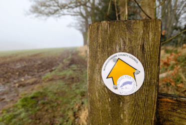 An Usk Valley Walk sticker on a footpath waymarker mounted to a wooden post in a farmer's field.