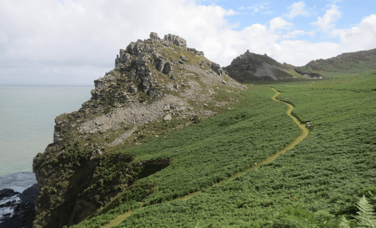 A narrow pedestrian footpath winds up the grassy flank of a huge rocky projection on the Exmoor coast, known as the Valley of Rocks.