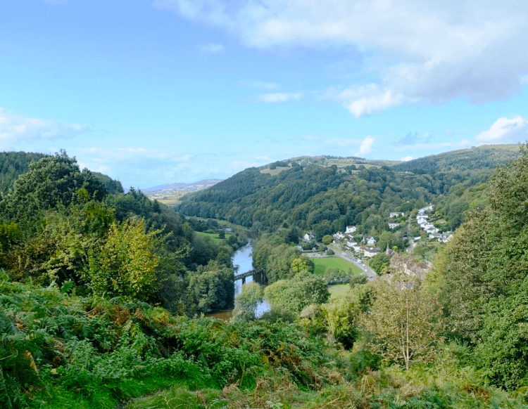 The view from high up: rounded hills covered with leafy trees surround a small town and a bridge crossing the River Wye.