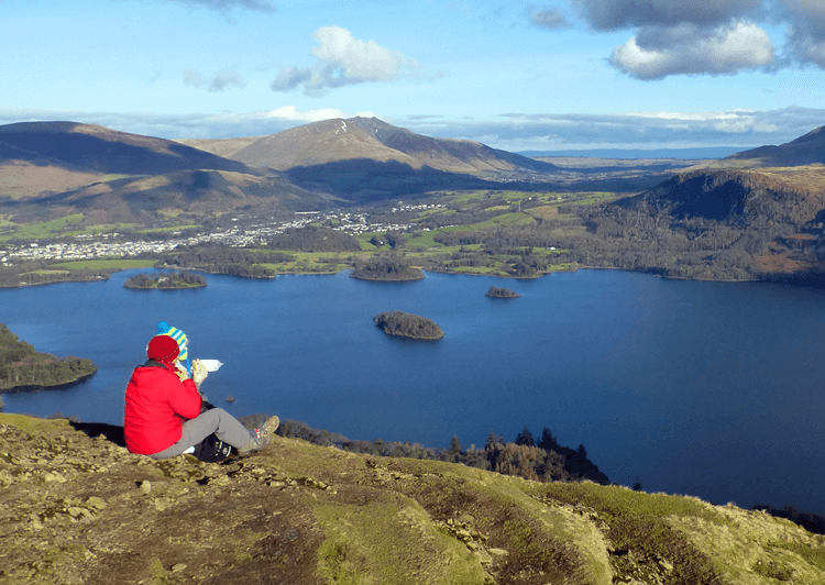 A pair of walkers in winter coats sit on Catbells overlooking a tarn as they eat lunch on the trail.