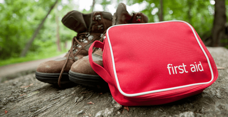 A red first aid kit sits propped up against a walker's boots in the middle of a forest. Presumably they're off treating their blisters just out of the shot.