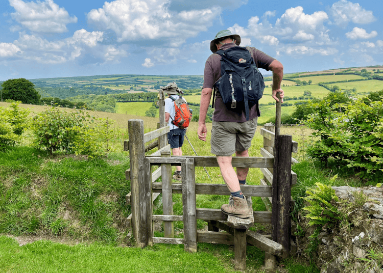 Walkers in summer hats and clothing clamber over a stile to continue through the British countryside.