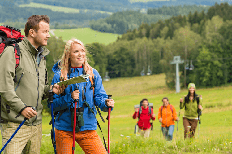 A group of hikers ascend a hill using walking poles. Two have already reached the top, and they use the time to orientate themselves with a paper map.