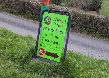 A lime green swinging sign sits beside the Usk Valley Walk footpath, advertising The Walnut Tree Village Shop & Cafe Takeaway.