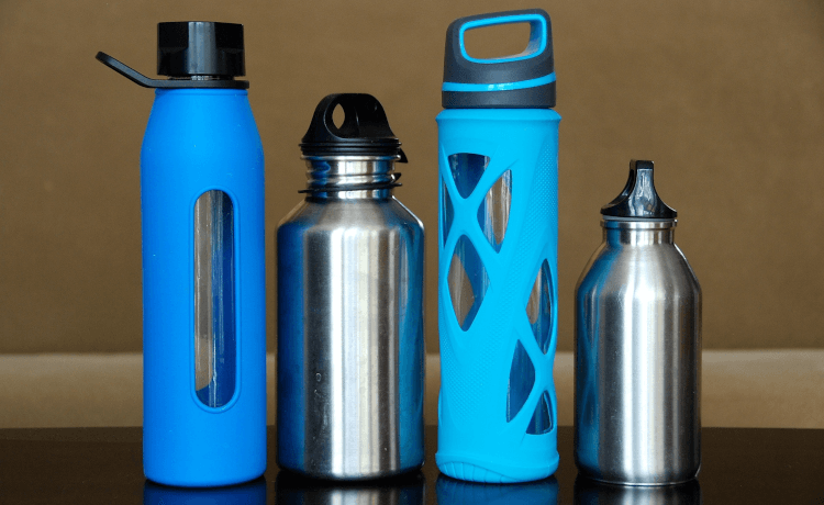 An array of reusable water bottles in glass, blue plastic and stainless steel, sit in a row on a table.