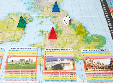 A close-up of the Who Knows Where boardgame, featuring four semi-transparent pyramid pieces and a di sitting on a map of the UK and Ireland with several cards laid beside them featuring famous places in the country.