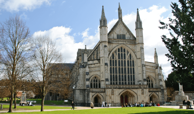 A head-on view of Winchester Cathedral, the start of the Pilgrims' Way. Surrounded by fine lawns and trees, the cathedral is built of pale stone, with an enormous arching set of windows, several ornate towers and supportive buttresses ranked down its sides.