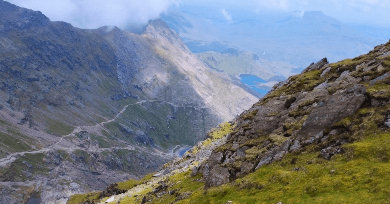 A view down over the lakes from Snowdon. Thin, winding dirt trails pick their way across the scree toward two expanses of blue water below the mountain.